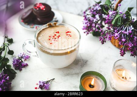 Dalgona Kaffee mit Schaum und Schokoladenchips in Form von Herzen, Schokoladenkekse, brennende Kerzen und frische, üppige lila-lila Blüten auf einem Licht Stockfoto