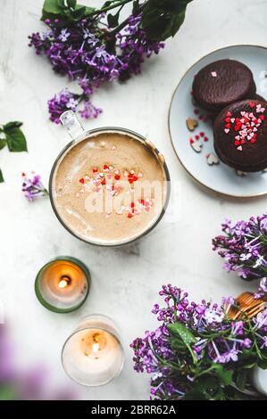 Dalgona Kaffee mit Schaum und Schokoladenchips in Form von Herzen, Schokoladenkekse, brennende Kerzen und frische, üppige lila-lila Blüten auf einem Licht Stockfoto