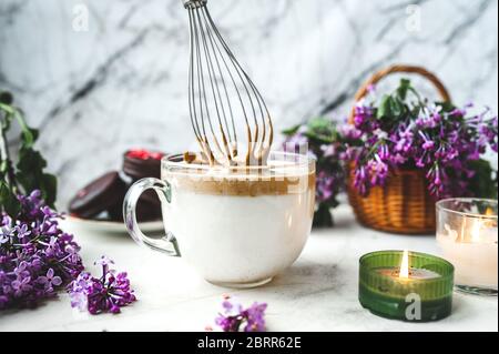 Metallkitchenbesen verquirlen Kaffeeschaum in einem großen Glas Tasse mit Milch auf einer weißen Marmoroberfläche mit einer brennenden Kerze, ein Weidenkorb mit einem üppigen b Stockfoto