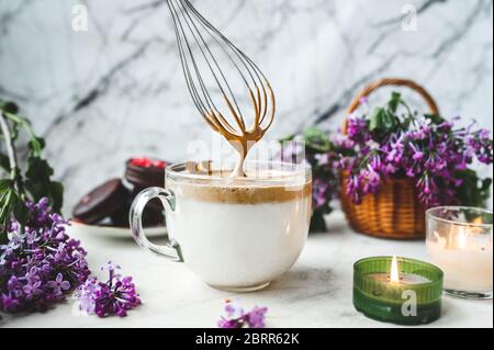 Metallkitchenbesen verquirlen Kaffeeschaum in einem großen Glas Tasse mit Milch auf einer weißen Marmoroberfläche mit einer brennenden Kerze, ein Weidenkorb mit einem üppigen b Stockfoto
