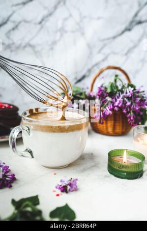 Metallkitchenbesen verquirlen Kaffeeschaum in einem großen Glas Tasse mit Milch auf einer weißen Marmoroberfläche mit einer brennenden Kerze, ein Weidenkorb mit einem üppigen b Stockfoto