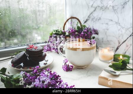Dalgona Kaffee mit Schaum, Schokoladenkekse, brennende Kerzen und frische, saftige lila-lila Blüten am nassen Fenster mit Tropfen und Tropfen vom Regen. Stockfoto
