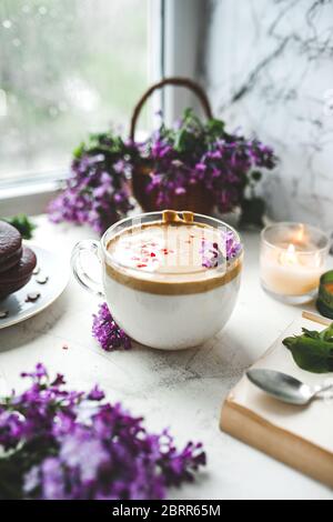 Dalgona Kaffee mit Schaum, Schokoladenkekse, brennende Kerzen und frische, saftige lila-lila Blüten am nassen Fenster mit Tropfen und Tropfen vom Regen. Stockfoto