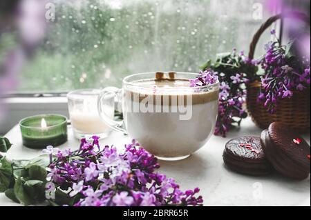 Dalgona Kaffee mit Schaum, Schokoladenkekse, brennende Kerzen und frische, saftige lila-lila Blüten am nassen Fenster mit Tropfen und Tropfen vom Regen. Stockfoto