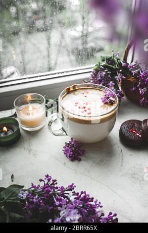 Dalgona Kaffee mit Schaum, Schokoladenkekse, brennende Kerzen und frische, saftige lila-lila Blüten am nassen Fenster mit Tropfen und Tropfen vom Regen. Stockfoto