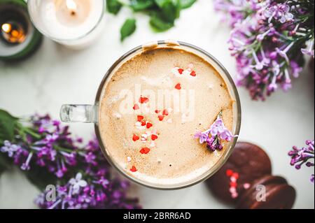 Dalgona Kaffee mit Schaum und Schokoladenchips in Form von Herzen, Schokoladenkekse, brennende Kerzen und frische, üppige lila-lila Blüten auf einem Licht Stockfoto