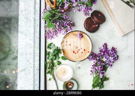 Dalgona Kaffee mit Schaum, Schokoladenkekse, brennende Kerzen und frische, saftige lila-lila Blüten am nassen Fenster mit Tropfen und Tropfen vom Regen. Stockfoto