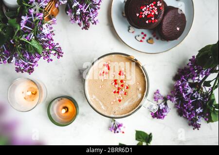 Dalgona Kaffee mit Schaum und Schokoladenchips in Form von Herzen, Schokoladenkekse, brennende Kerzen und frische, üppige lila-lila Blüten auf einem Licht Stockfoto