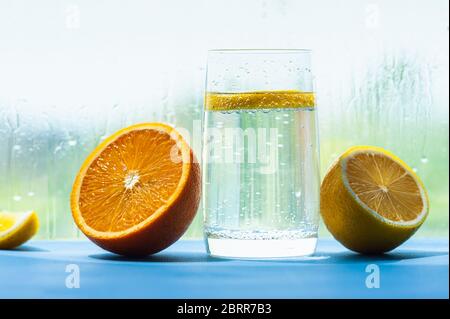 Transparentes Glas mit gasförmigem Wasser und Blasen und einer runden Scheibe Zitrone im Inneren, halb saftig und reif Orange und Zitrone daneben, Glas mit Tropfen Stockfoto