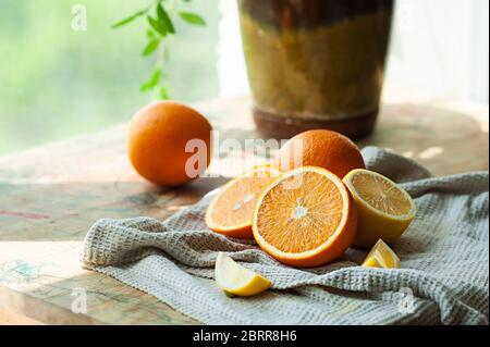 Rustikales Frühstück. Reife saftige ganze Orangen und Hälften auf einem baumwollgrauen Waffeltuch auf einem Hintergrund einer grünen Blume in einem Topf nahe dem Fenster. Vitami Stockfoto