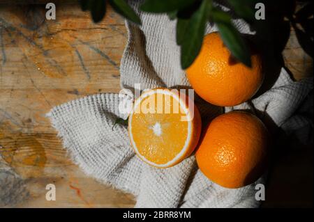 Rustikales Frühstück. Reife saftige ganze Orangen und Hälften auf einem baumwollgrauen Waffeltuch auf einem Hintergrund einer grünen Blume in einem Topf nahe dem Fenster. Vitami Stockfoto