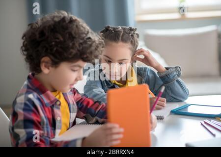 Lockiger Junge und dunkelhaarige Mädchen sitzen am Tisch mit Tabletten, ihre Aufgaben zu tun, beteiligt zu sein Stockfoto