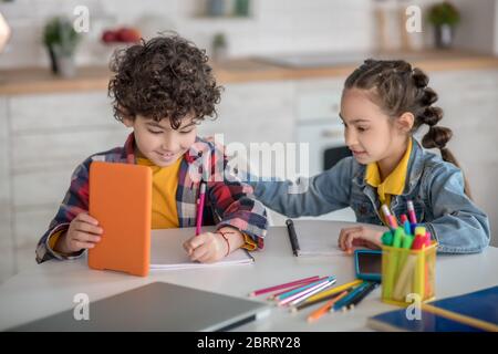Lockiger Junge und dunkelhaarige Mädchen sitzt am Tisch mit Tabletten, Mädchen, die ihre Hand auf junge Schulter Stockfoto