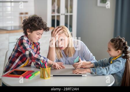 Müde blonde Frau sitzt am Tisch, hält ihren Kopf, lockigen Jungen und dunkelhaarige Mädchen kämpfen über Laptop Stockfoto