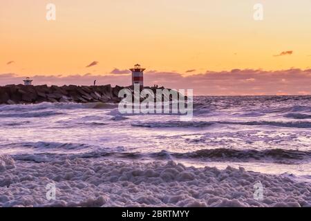 Rot-weiß gestreifter Leuchtturm bei einem lebhaften Sonnenuntergang. Wasser trifft die schwarzen Felsbrocken in Wellen. Dramatischer Himmel in goldener Farbe. Scheveningen, die Neth Stockfoto