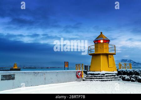 Leuchtturm am Eingang zum Alten Hafen, Reykjavik, Island. Blaue Stunde, die im Winter bei Sonnenaufgang geschossen wurde, mit gelben Leuchtfeuer und Schnee auf dem Boden. Stockfoto