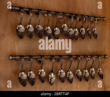 Zwei Reihen von glänzenden Edelstahl Löffel hängen an Geländern der braunen Wand in der Nähe von Fenster. Polierte reflektierende Oberfläche des Bestecks Stockfoto