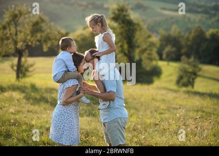 Junge Familie mit zwei kleinen Kindern, die bei Sonnenuntergang auf der Wiese im Freien stehen. Stockfoto