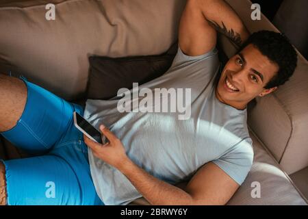 Draufsicht auf glücklich gemischt Rennen Mann chillen und mit Smartphone auf dem Sofa während der Selbstisolierung Stockfoto