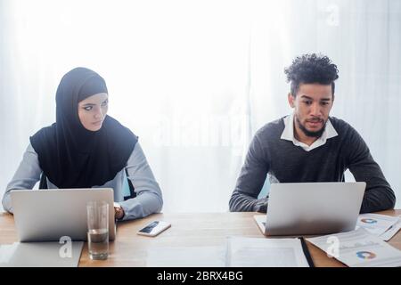 Selektiver Fokus der muslimischen Geschäftsfrau, die sich während ihrer Arbeit im Büro einen afroamerikanischen Kollegen anschaut Stockfoto