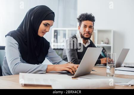Selektiver Fokus arabischer Geschäftsfrau, die Laptop in der Nähe afroamerikanischer Kollegen im Büro benutzt Stockfoto