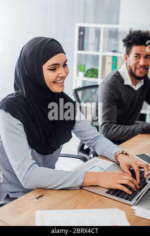 Selektiver Fokus der lächelnden arabischen Geschäftsfrau mit Laptop in der Nähe von afroamerikanischen Kollegen im Büro Stockfoto