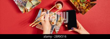 Panoramabild der Frau, die Essstäbchen mit gedämpftem Brötchen und Smartphone mit leerem Bildschirm in der Nähe von leckeren chinesischen Speisen in Take-away-Boxen auf rot hält Stockfoto