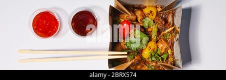 Panorama-Aufnahme von Saucen und Essstäbchen in der Nähe von Take-away-Box mit zubereitetem Schweinefleisch und Gemüse auf weiß Stockfoto
