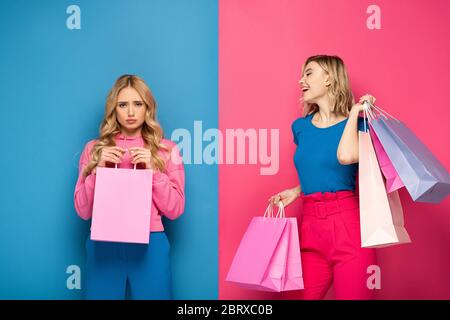 Lächelndes Mädchen mit Einkaufstaschen, die beleidigte Schwester auf rosa und blauem Hintergrund ansieht Stockfoto