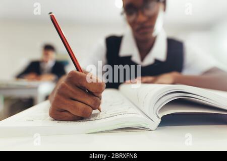 Nahaufnahme eines Teenagers, das in ihr Lehrbuch schreibt, während es im Klassenzimmer am Schreibtisch sitzt. Schüler der High School konzentriert beim Schreiben in einem Buch, Focu Stockfoto