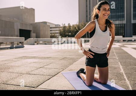 Fit Frau kniet auf Trainingsmatte im Freien. Frauen in Sportbekleidung gehen Yoga in der Stadt. Stockfoto