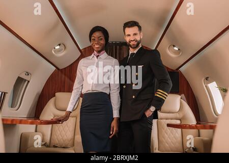 Schöner, positiver Pilot, der mit der Hand in der Tasche in der Nähe der lächelnden afroamerikanischen Stewardess im Privatjet steht Stockfoto