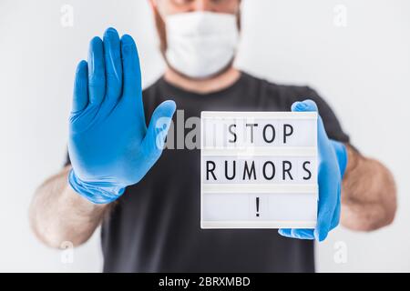 Fake News Infosemics during Covid-19 Pandemie Concept. Mann trägt Schutzmaske und medizinische Handschuhe an den Händen halten Leuchtkasten mit Text Stop Rumors. Stockfoto