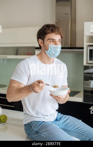 Schöner kranker Mann in medizinischen Maske hält Schüssel mit Cornflakes auf Küche auf Selbstisolierung Stockfoto