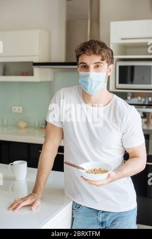 Kranker Mann in medizinischer Maske hält Schüssel mit Cornflakes auf Küche auf Selbstisolierung Stockfoto