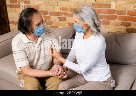 Reife Frau in medizinischen Maske hält digitales Thermometer in der Nähe Ehemann Stockfoto
