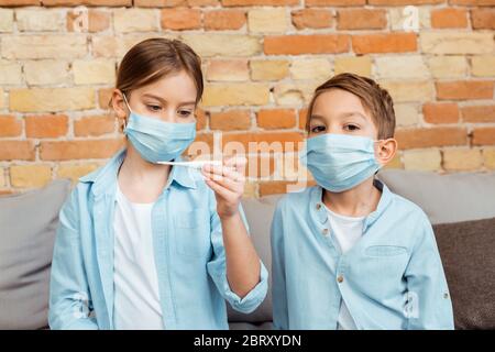 Kranke Kind in medizinischen Maske hält digitales Thermometer in der Nähe Bruder Stockfoto