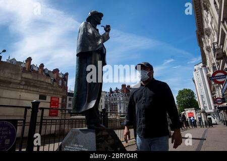 Ein Mann in einer Gesichtsmaske geht am Sherlock Holmes Day vor der U-Bahnstation Baker Street an der Sherlock Holmes Statue vorbei, nachdem Maßnahmen eingeführt wurden, um das Land aus der Blockierung zu bringen. Stockfoto