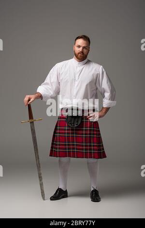 Schottischer Rotschopf-Mann in rotem Kilt mit Schwert auf grauem Hintergrund Stockfoto