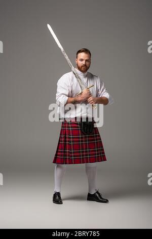 Schottischer Rotschopf-Mann in rotem Kilt mit Schwert auf grauem Hintergrund Stockfoto