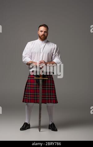 Schottischer Rotschopf-Mann in rotem Kilt mit Schwert auf grauem Hintergrund Stockfoto