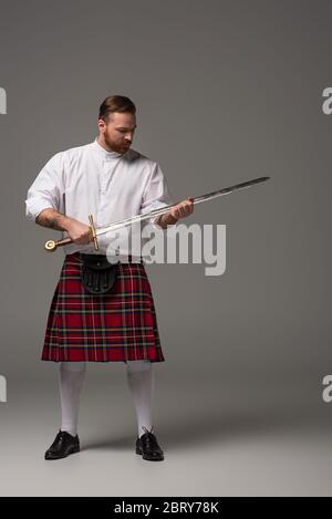 Schottischer Rotschopf-Mann in rotem Kilt mit Schwert auf grauem Hintergrund Stockfoto