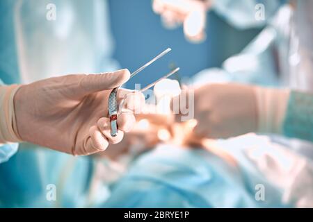 Arzthand hält eine Pinzette im Operationssaal. Chirurgische Instrumente in der Hand des Arztes auf dem Hintergrund des Operationssaal mit Ärzten Stockfoto