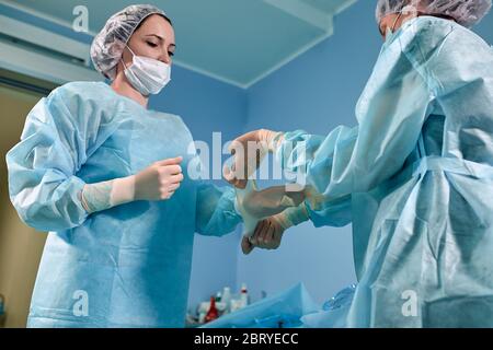 Ein Team von Chirurgen bereitet sich auf die Operation vor. Chirurgen tragen sterile Kleidung vor der Operation mit Hilfe von Krankenschwestern, sterile Kleider, Handschuhe, Masken, in Stockfoto