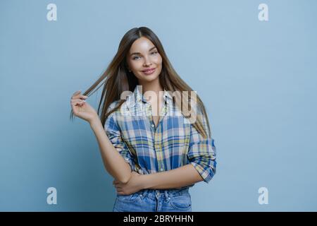 Halbe Länge Schuss von hübschen jungen Frau mit dunklen Haaren, trägt stilvolle Kleidung, hat europäische Erscheinung, gilt Make-up, posiert im Studio gegen blaue BA Stockfoto