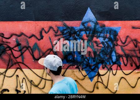 Berlin, Deutschland. Mai 2020. Ein junger Mann geht in der East Side Gallery an dem zerstörten Wandgemälde des Künstlers Günther Schäfer vorbei. Das Wandbild Vaterland, das die schwarz-rot-goldene deutsche Flagge mit einem blauen Davidstern zeigt, wurde von Unbekannten mit schwarzen Graffiti beschmiert. Kredit: Carsten Koall/dpa/Alamy Live News Stockfoto