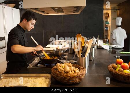 Männlicher Kochmütze, der beim Herd in der Küche steht, das Essen im Wok mit dem Holzlöffel rührend. Stockfoto