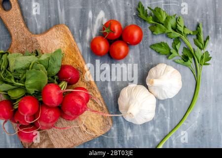 Hochwinkelaufnahme von frisch gepflückten Radieschen, Knoblauch und flacher Petersilie und Holzhackbrett auf grauem Marmor. Stockfoto