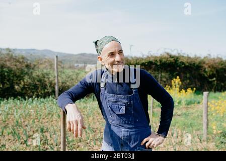 Lächelnder Mann mit Latzhose und Bandana, der im Gemüsegarten steht und sich an Holzpfosten lehnt. Stockfoto