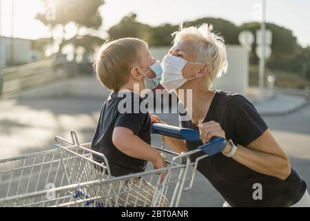 Mutter spielt freudig mit Kind im Einkaufswagen sitzen. Beide tragen eine schützende Gesichtsmaske, bevor sie in einen Supermarkt gehen. Stockfoto